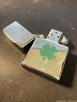 Photo of the silver-tone Zippo lighter opened to show the windproof chimney and flint wheel. The front of the lighter has a green four leaf clover and the word 'LUCKY'.