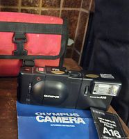 Olympus 35mm film camera with built-in flash, A16 flash unit, user manuals, and red carrying case displayed on wooden surface.