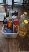 Photo showing various cleaning and bug repellent sprays and liquids in a white plastic container on the floor alongside two large Pine-Sol cleaning bottles.