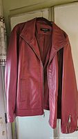 Front view of reddish faux leather jacket hanging, showing full zip front, side pockets, collar, and general style and color of the jacket.