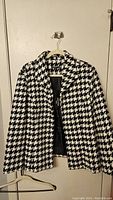 Front view of a black and cream houndstooth patterned women's jacket on a hanger against a door, showing lapel collar, button closure, and long sleeves.