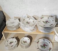 Photo of stacked china plates and bowls showing pink rose floral pattern, stacks on shelving.
