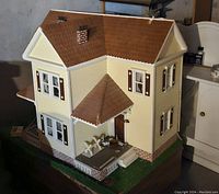 Front and side view of Victorian style dollhouse showing detailed exterior including porch, windows, shutters, chimney, and shingled roof. Surrounded by some small furniture and accessories on green base.