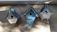 Front view of three wooden birdhouses with peaked roofs and hanging wires, painted blue and white with weathering visible.