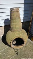 Full front view of the chiminea showing open mouth, round body with brick pattern, mossy discoloration, and metal stand legs visible at base.