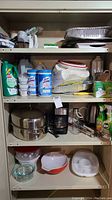 Four shelves filled with kitchen items and cleaning products including metal roasting pan, coffee maker, cleaning sprays, paper products, and various bowls.