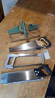 Three different saws placed next to miter box on wooden table showing variety and condition.
