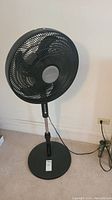 Tall black standing fan with circular grille and five blades, adjustable height, and control buttons on the neck, shown plugged into a wall outlet.
