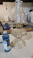 Full view of vintage glass oil lamp with chimney and bottle of blue lamp oil next to it on table in basement.