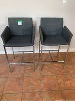 Front view of two grey linen-look upholstered counter height stools with chrome bases on a tile floor.