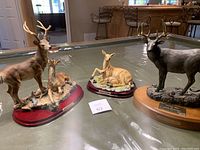 Three deer figurines on a green table: two natural brown and tan colored deer on red bases, and one dark grey on wooden base with plaque.