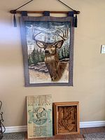 Full view of the three deer-themed wall decor pieces hanging on a beige wall: buck tapestry above, wooden sign and relief plaque below.