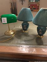 Photo of all three lamps on a pool table showing the green banker’s lamp and two green ceramic night table lamps with floral designs and pleated shades.