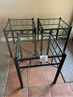 Three of the four dark green metal framed end tables with clear square glass tops arranged closely together on a tile floor.
