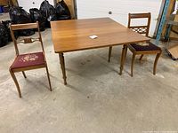 Antique solid wood drop-leaf table with two vintage wooden chairs with needlepoint seats, displayed in garage surroundings on concrete floor.