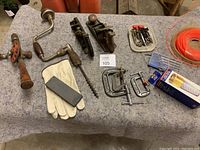 Overview of antique hand planes, hand drills, Allen keys, weed whipper string, clamps, cutter sharpening block with gloves, and foot valve on gray cloth.