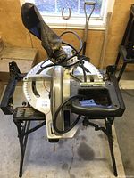 Front angled view of Delta sliding compound miter saw mounted on black foldable stand, showing handle, blade guard, and dust collection bag, placed on concrete floor near window.