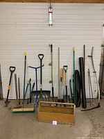 Full view of all garden tools arranged upright against a wall, showing shovels, push brooms, bolt cutters, wooden toolbox, scythe, and garden claw.