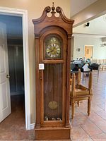 Full vertical view of the solid oak grandfather clock with carved pediment and finial, showing full height and presence in a tile floor room.