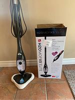 Upright black steam mop with triangular mop head and cleaning pad beside open box showing product branding and features.