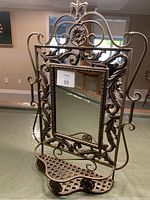 Full frontal view of the iron mirror showcasing the intricate floral and scrollwork around the mirror and the attached bottom shelf with curled iron legs.