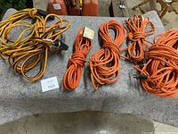Six outdoor extension cords of various lengths and colors arranged on a gray surface. Mostly orange color with black accents and visible 3-prong plugs.