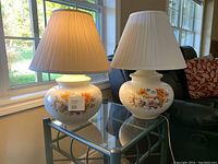 Pair of white ceramic lamps on glass table by window, each with colorful floral decals and pleated white shades.