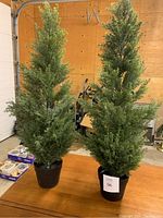 Full view of the pair of faux evergreen trees standing on a wooden table in a garage setting, showing full height and pots.