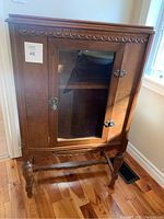 Front view of small wooden cabinet with glass door and brass hardware. Shows carved top trim and sculpted legs.