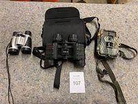 Photo of the lot containing a small pair of Vivitar binoculars, Tasco binoculars with a carrying case, and a camouflage Bushnell Trophy Camera with strap on a grey countertop.