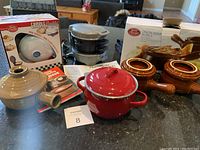 Full view of lot showing all kitchen items including boxed Betty Crocker mini griddle, stack of metal mini griddles, ceramic stoneware cooker, boxed French onion soup bowls, and small red pot with lid.