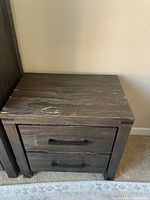 Photo of espresso-colored solid wood nightstand showing top surface and two drawer fronts with black handles, visible wear and scratches on the top and sides.