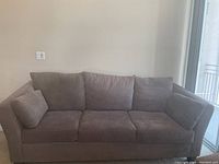 Grey three-seater fabric sofa with three seat cushions, three back cushions, and two side cushions. Positioned against a plain wall near a window.