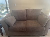 Photo of a two-seater grey sofa with cushioned back and seat, showing the left and right armrests with additional side cushions, placed indoors near a balcony window.