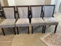 Photo of four Parson-style dining chairs arranged side by side showing grey fabric upholstery with some wear marks and solid dark wooden legs, set on a carpeted floor.