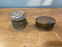 Two small handmade boxes on a wooden surface, one round metal box with a lid and one oval wooden box.