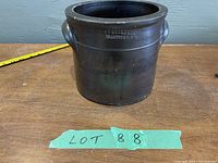 Front view of the antique brown crock on wooden surface with measuring tape showing height approximately 8 inches. The crock has two handles and embossed maker's mark on the side.