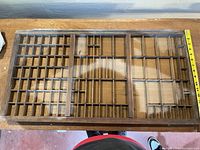 Top-down view of wooden display board showing multiple small compartments arranged in a grid pattern, measuring about 16 inches wide as verified by measuring tape.