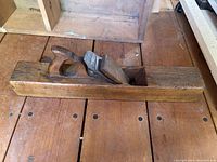 Long antique wooden hand plane placed on wooden floor, showing the top, handle, and body clearly.