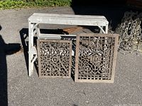 Two antique Victorian cast iron cold air return grates displayed leaning against a white foldable frame on asphalt.