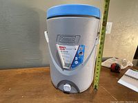Full view of grey Coleman beverage cooler with blue lid and white handle, next to a tape measure showing height around 15 inches.