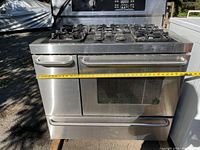 Front view of stainless steel gas stove showing six gas burners on top, oven door with window, warming oven door on left side, and tape measure indicating width about 41 inches