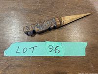 Wooden letter opener with detailed hand carved indigenous design placed on a wooden surface next to a green label marked 'LOT 96'.