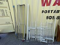 Various metal wire shelving units and components including tall and smaller wire shelves, support bars, and frame elements displayed outdoors.