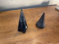 Two metal wood splitting wedges standing on a wooden surface against a wall, showing their pointed shapes and blackened surfaces with wear and rust.