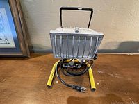 Front view of the industrial work light showing protective grill, handle, yellow base, and power cord
