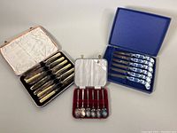 Three vintage cutlery sets displayed in their original cases: porcelain steak cutters with blue onion pattern in blue case, silver plated coffee spoons in red case, and fish fork/cutter set with cream celluloid handles in cream and black case.