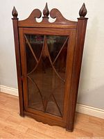 Front view of the antique cherry wood corner cupboard showing the glass door with wooden muntin design and finial decorations.