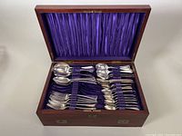 Open wooden chest displaying arranged flatware pieces including spoons, forks, and knives on purple fabric lining.