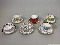 Overview of six assorted teacup and saucer sets arranged in two rows, featuring different floral and thematic designs in fine china with gold trim
